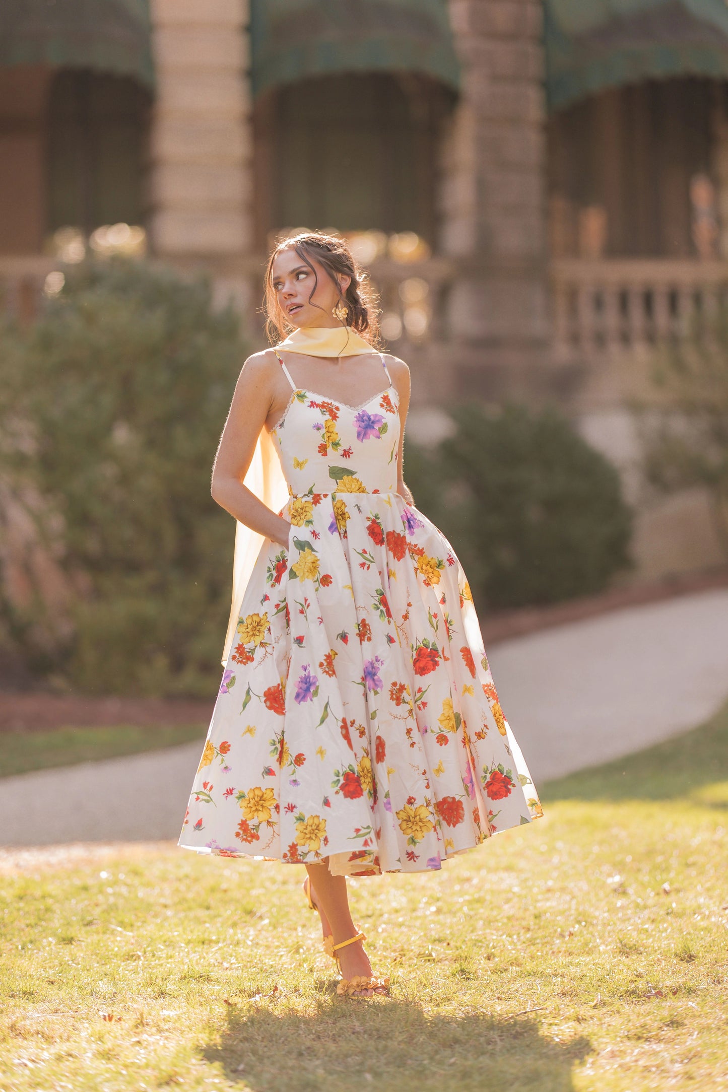 The image shows a woman in a white dress walking through a field of grass, with a building and trees in the background. She is wearing a pair of footwear, suggesting she is ready to take on the world.