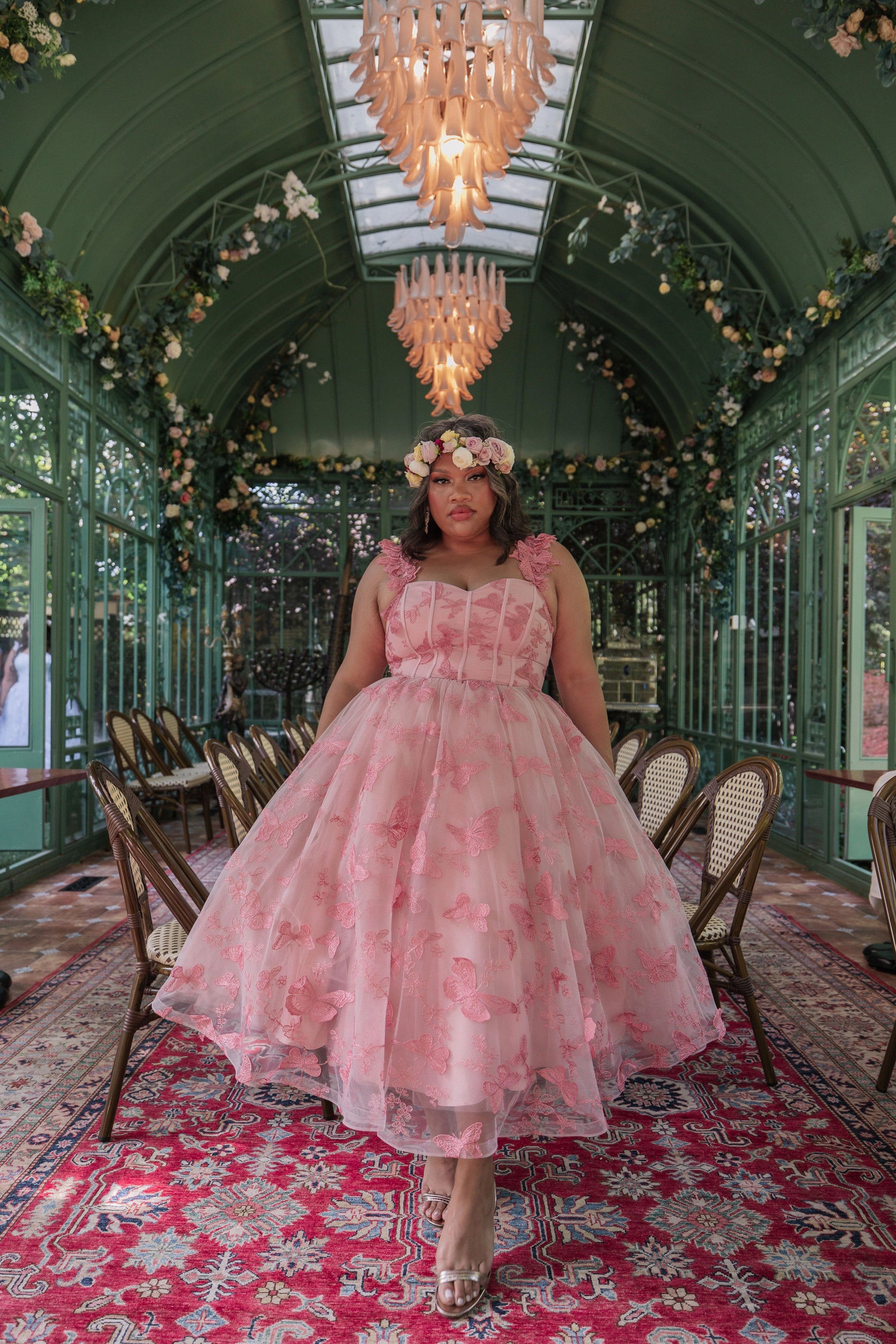 The image shows a woman in a pink dress standing on a red carpet in a room with chairs, a chandelier, and a glass wall in the background.