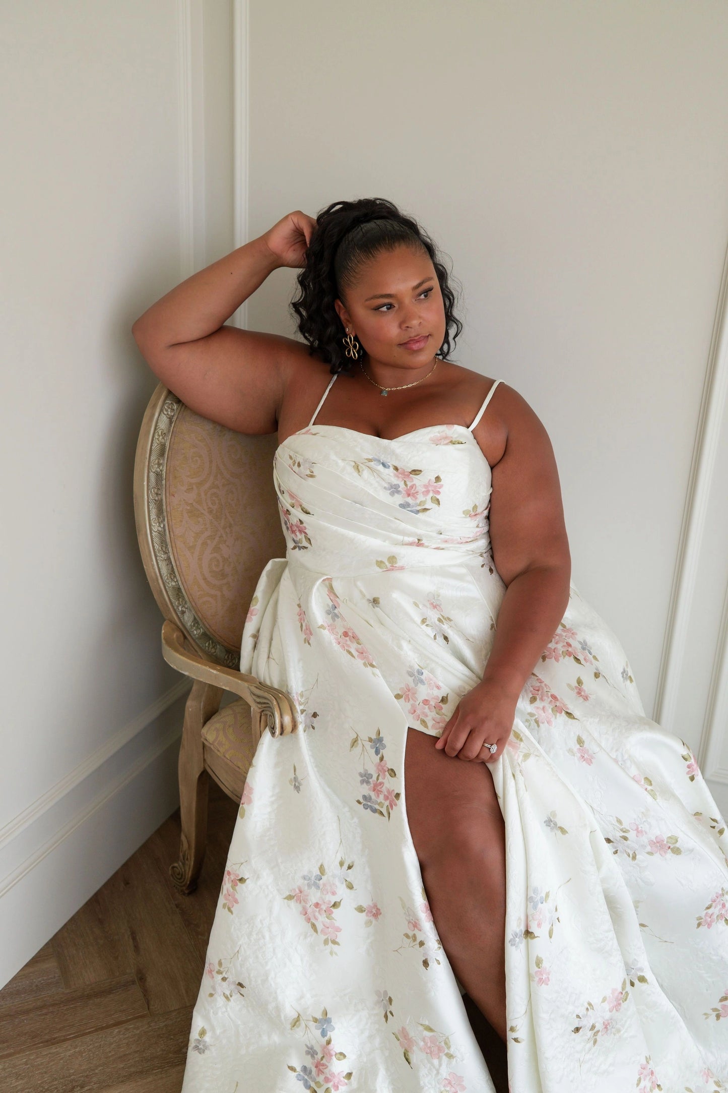 The image shows a woman in a white dress sitting on a chair in front of a wall. She is wearing a white frock and her hair is styled in an elegant updo. Her dress is long and flowy, with a high