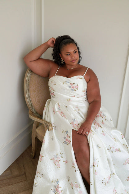 The image shows a woman in a white dress sitting on a chair in front of a wall. She is wearing a white frock and her hair is styled in an elegant updo. Her dress is long and flowy, with a high