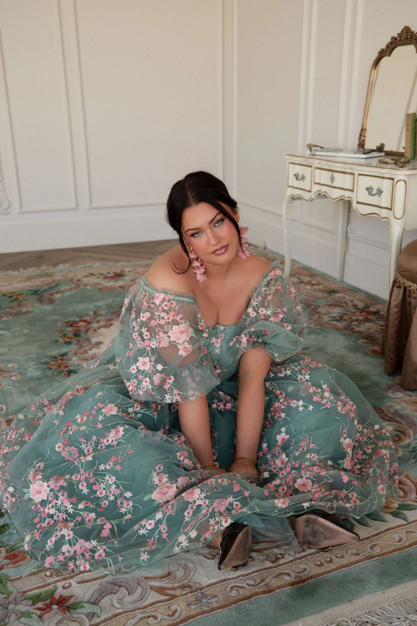 The image shows a woman in a green dress sitting on the floor in front of a mirror, with a table to her right and a wall in the background.