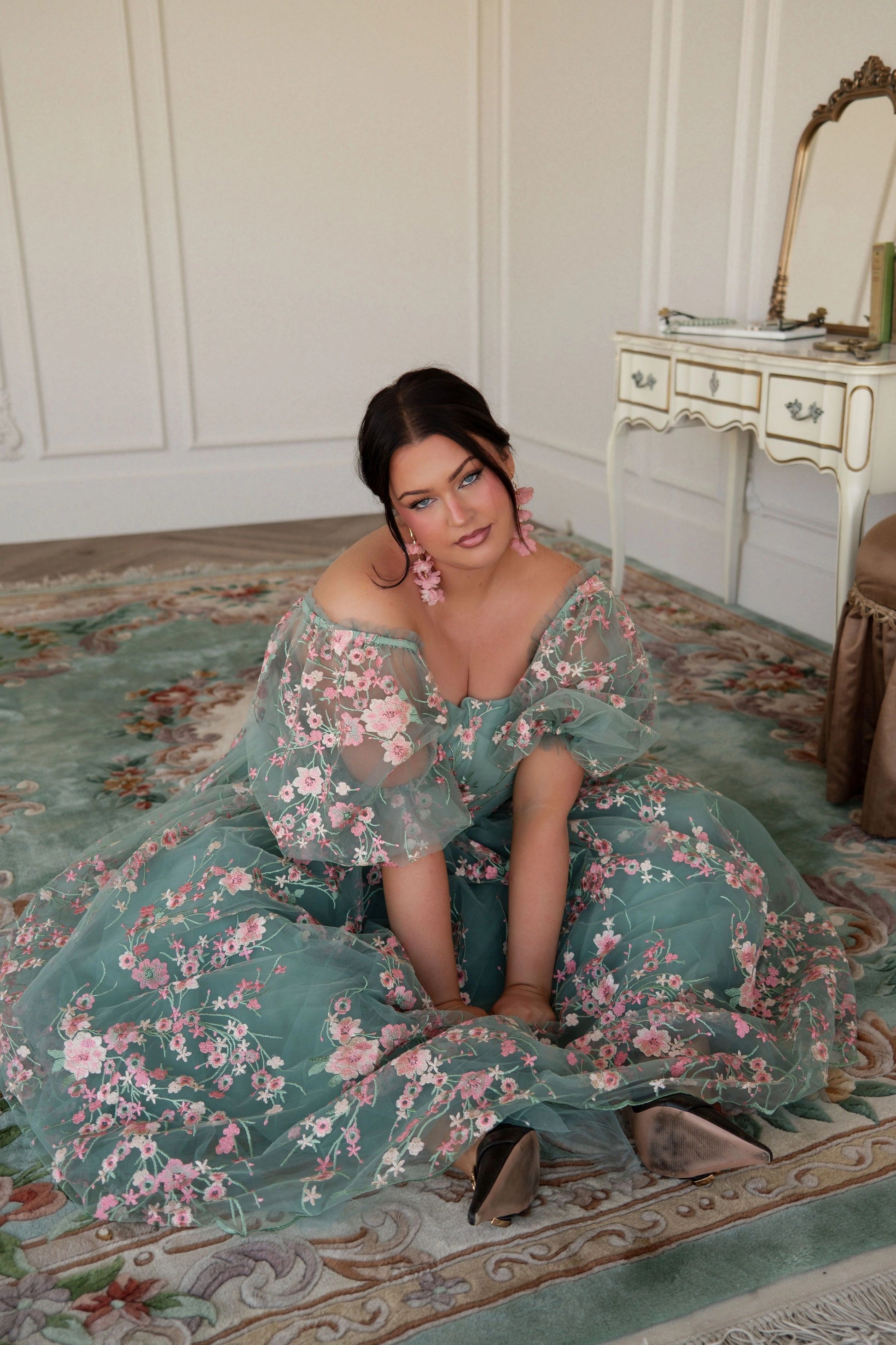 The image shows a woman in a green dress sitting on the floor in front of a mirror, with a table to her right and a wall in the background.