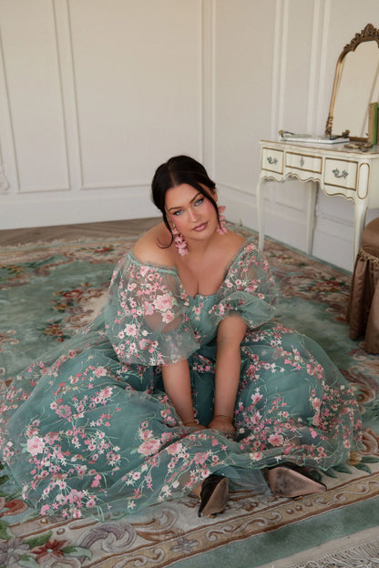 The image shows a woman in a green dress sitting on the floor in front of a mirror, with a table to her right and a wall in the background.