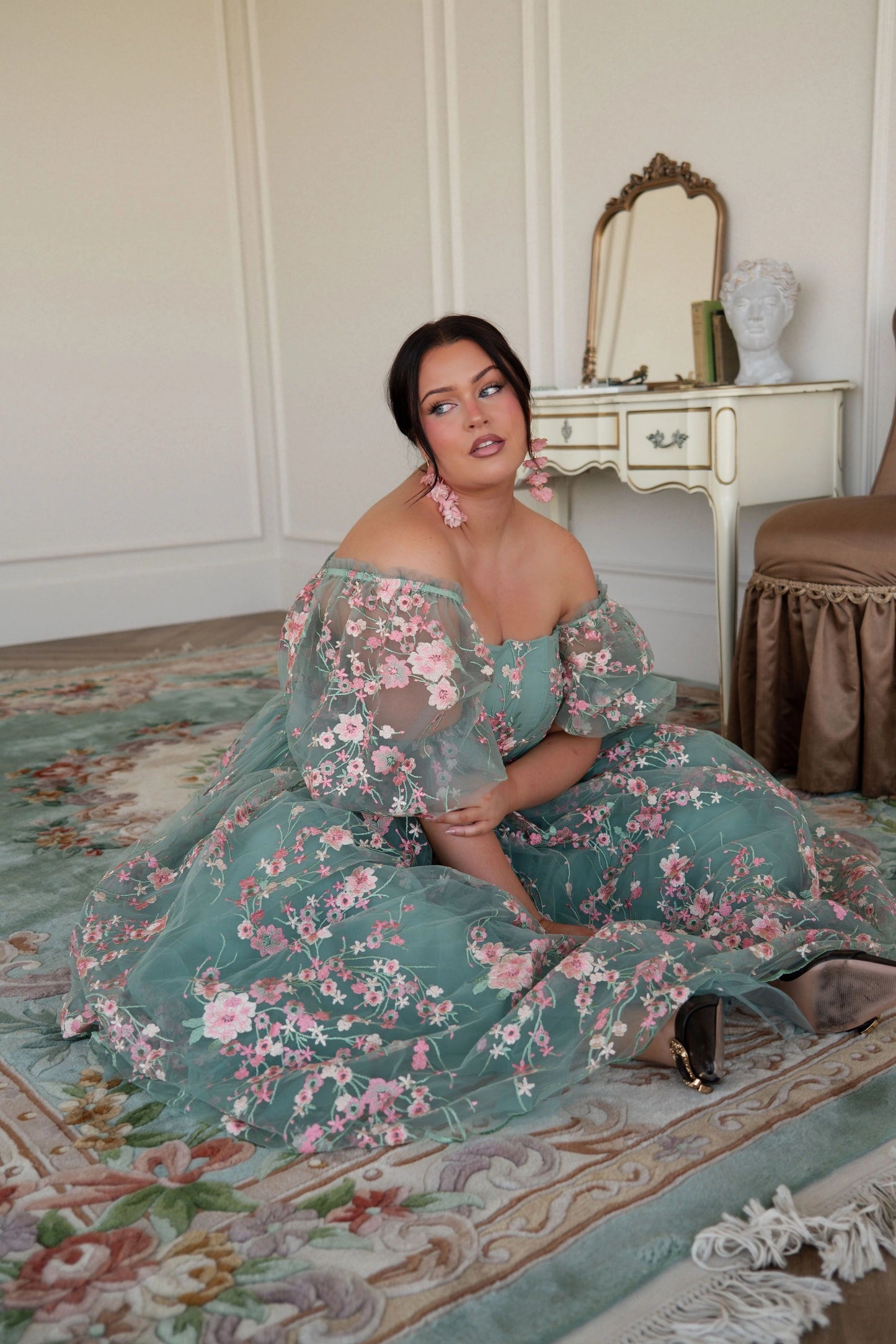 The image shows a woman in a green dress sitting on the floor in front of a mirror, with a chair and a table in the background. The wall behind her is visible, and there is text at the bottom of the
