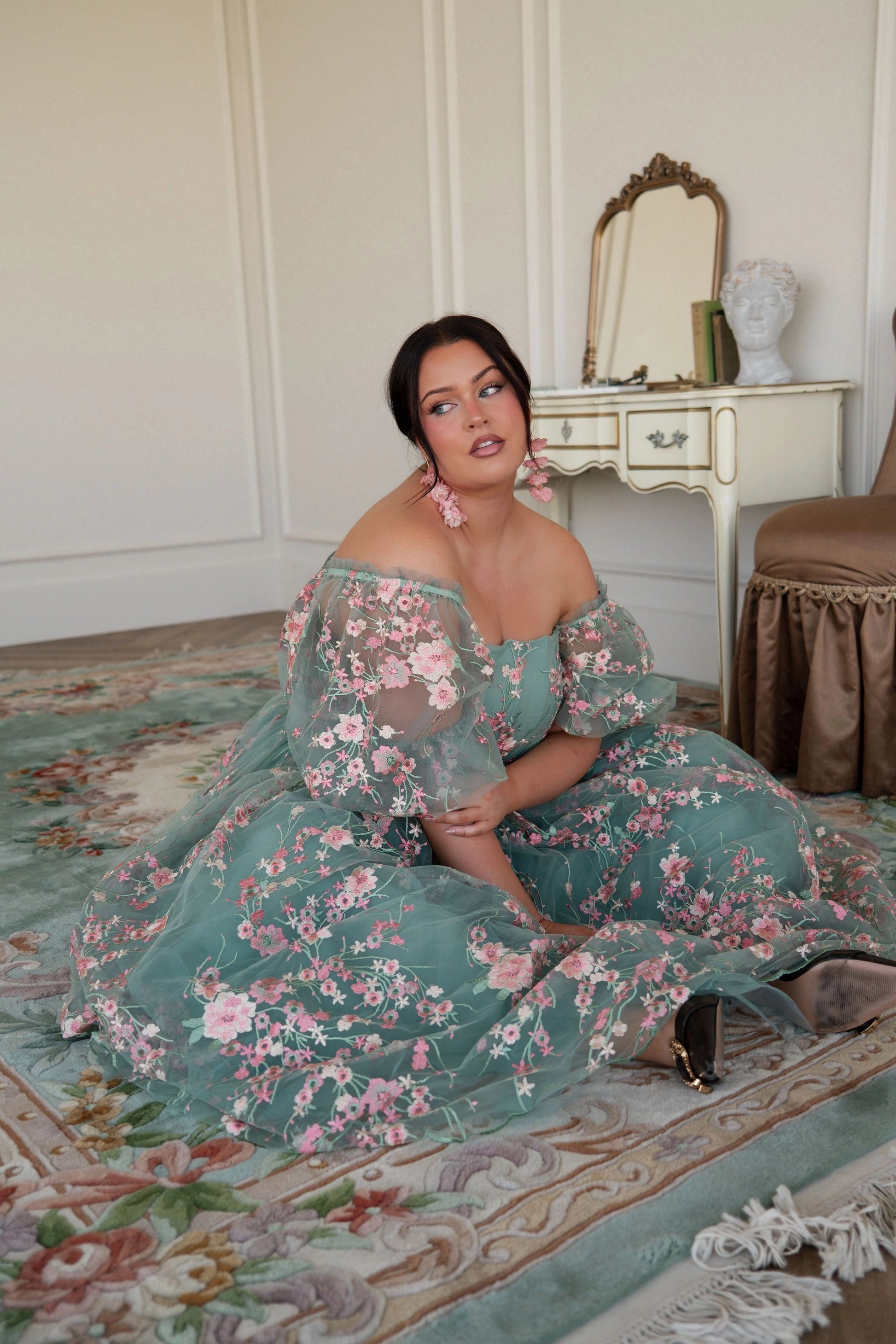 The image shows a woman in a green dress sitting on the floor in front of a mirror, with a chair and a table in the background. The wall behind her is visible, and there is text at the bottom of the