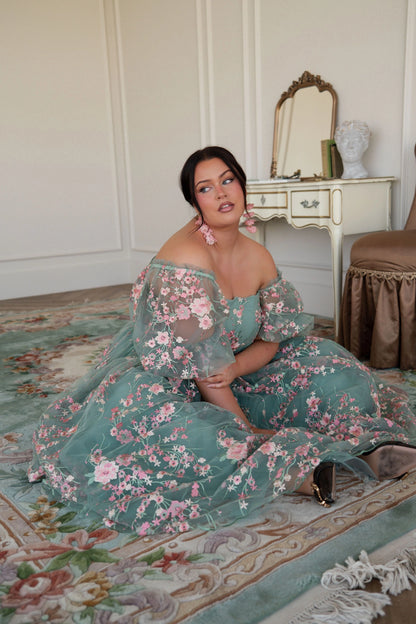The image shows a woman in a green dress sitting on the floor in front of a mirror, with a chair and a table in the background. The wall behind her is visible, and there is text at the bottom of the