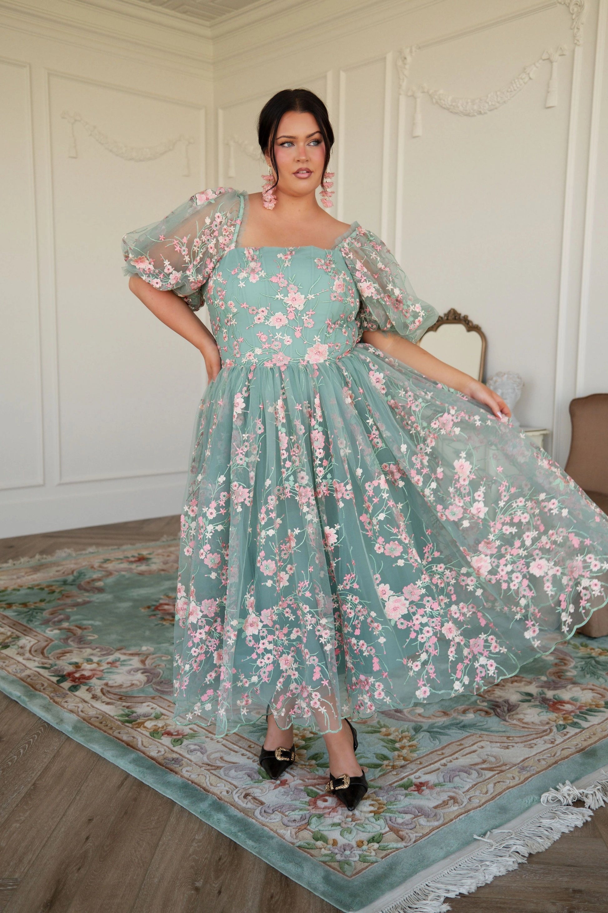 The image shows a woman in a green dress standing on a carpeted floor, with a chair and a wall in the background. She is wearing a dress with a high neckline and long sleeves, and her hair is styled