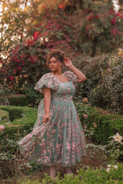 Woman in green floral dress with pink flowers and ruffled sleeves poses in garden with bushes and flowers.