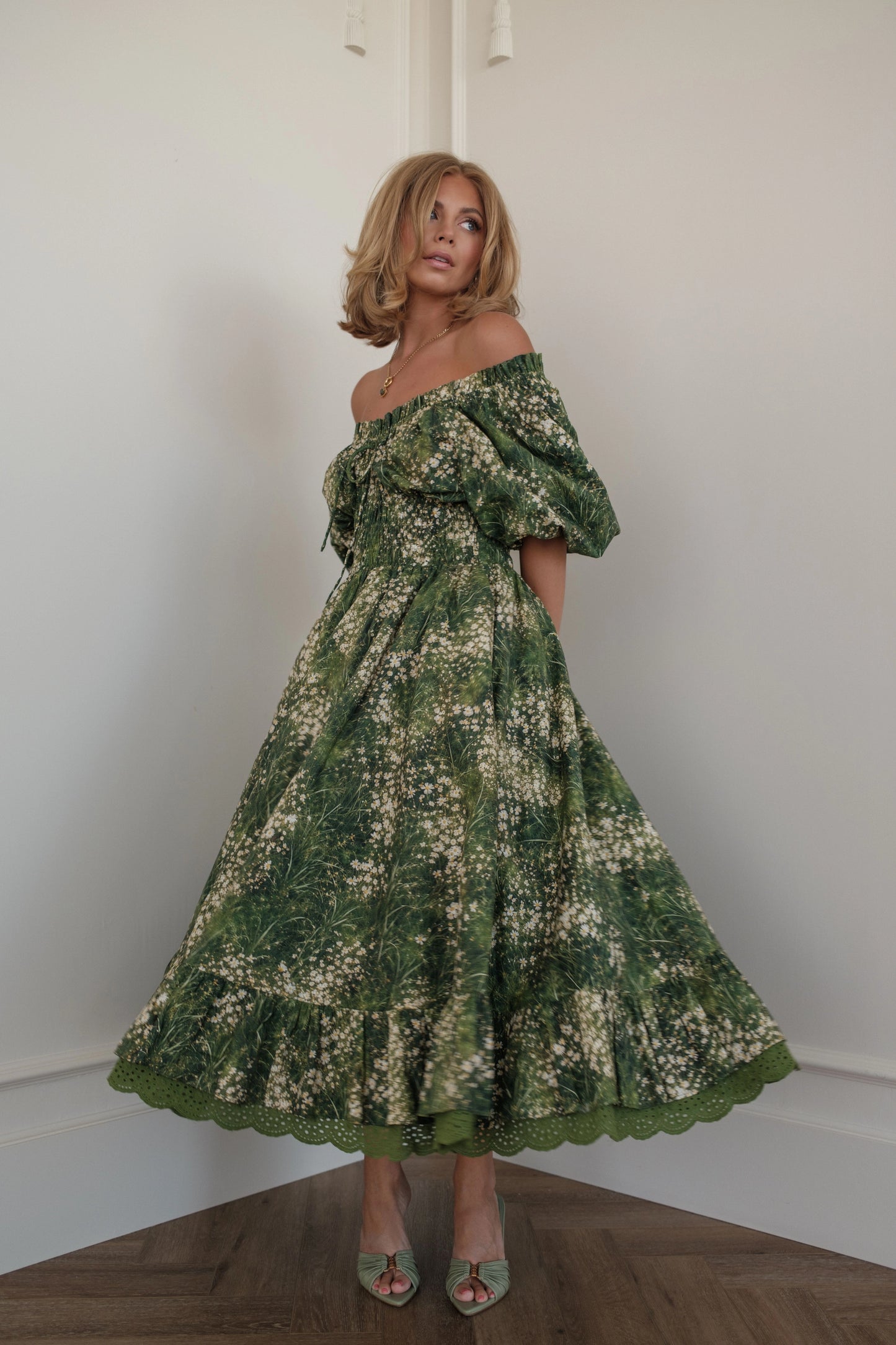 Woman in green floral off-the-shoulder dress and heels poses in corner of white room.