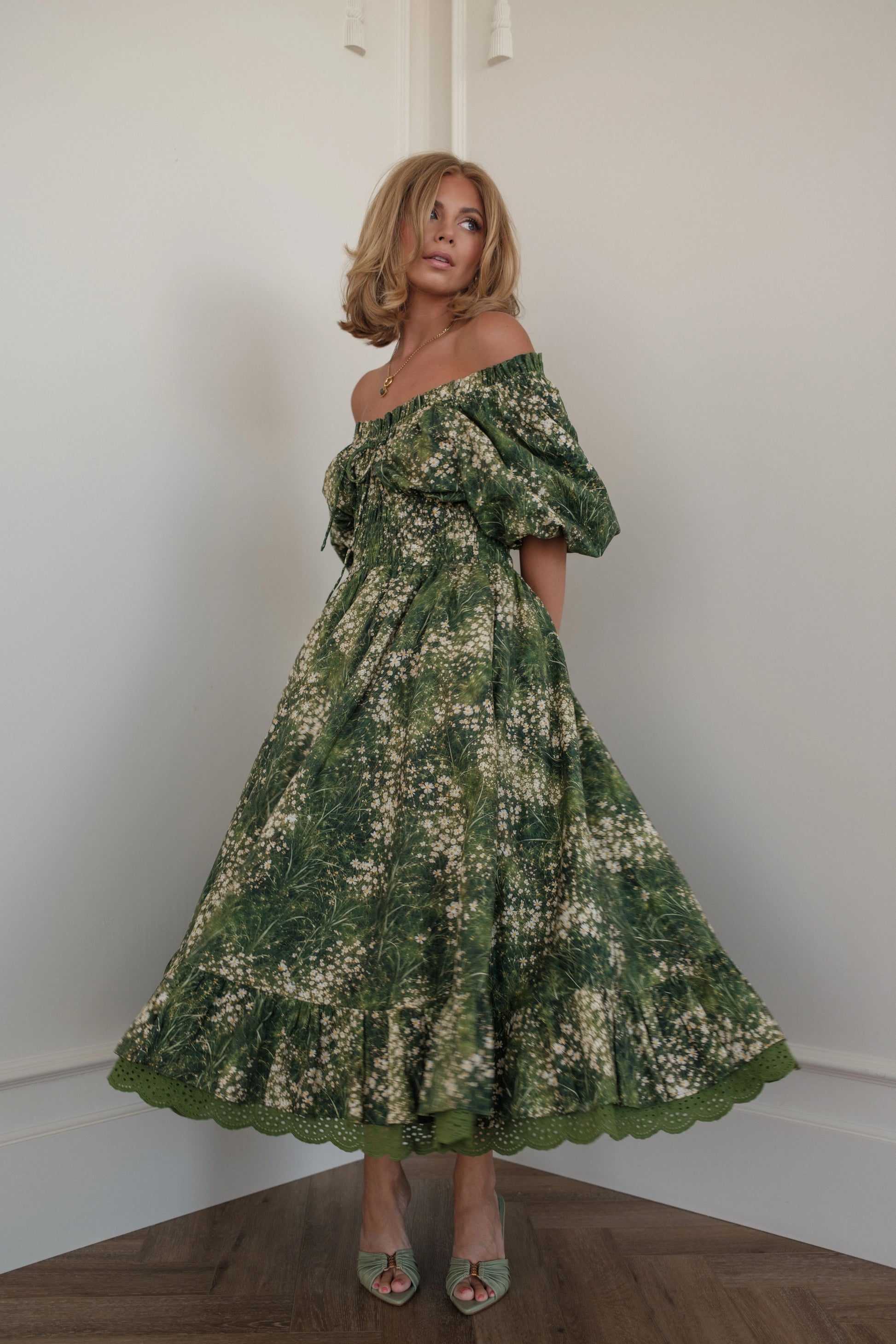 Woman in green floral off-the-shoulder dress and heels poses in corner of white room.