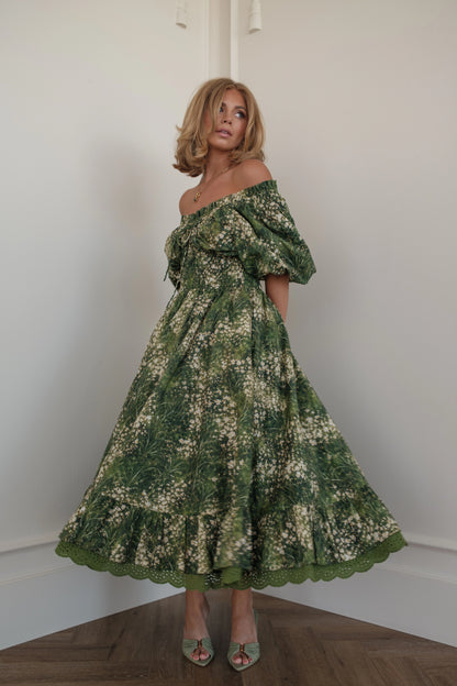 Woman in green floral off-the-shoulder dress and heels poses in corner of white room.