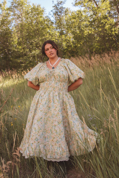 The image shows a woman in a dress standing in a field of tall grass, surrounded by trees and a clear blue sky.