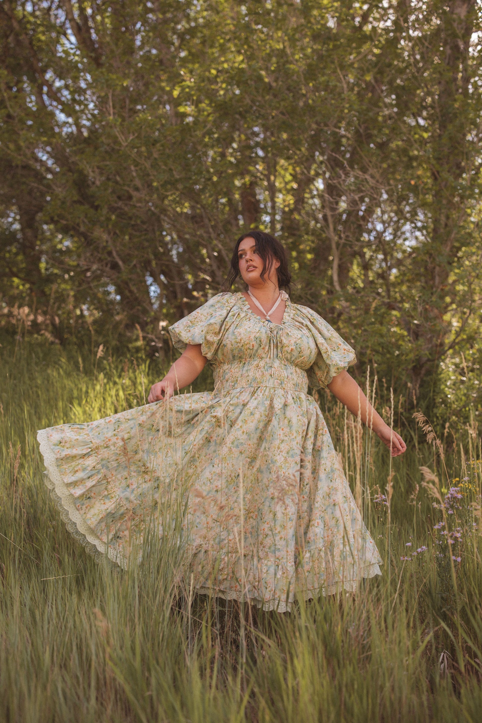 The image shows a woman in a long dress walking through tall grass, with trees in the background and a clear blue sky above.