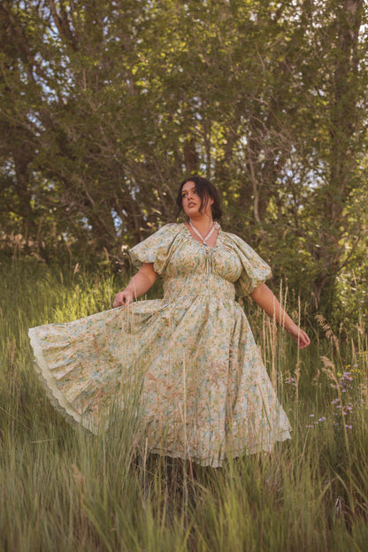 The image shows a woman in a long dress walking through tall grass, with trees in the background and a clear blue sky above.