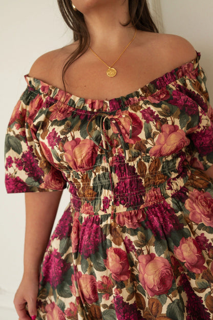 The image shows a woman wearing a long dress with a floral print, standing in front of a wall. The dress is a light color with a pattern of small flowers in shades of pink, purple, and white. The