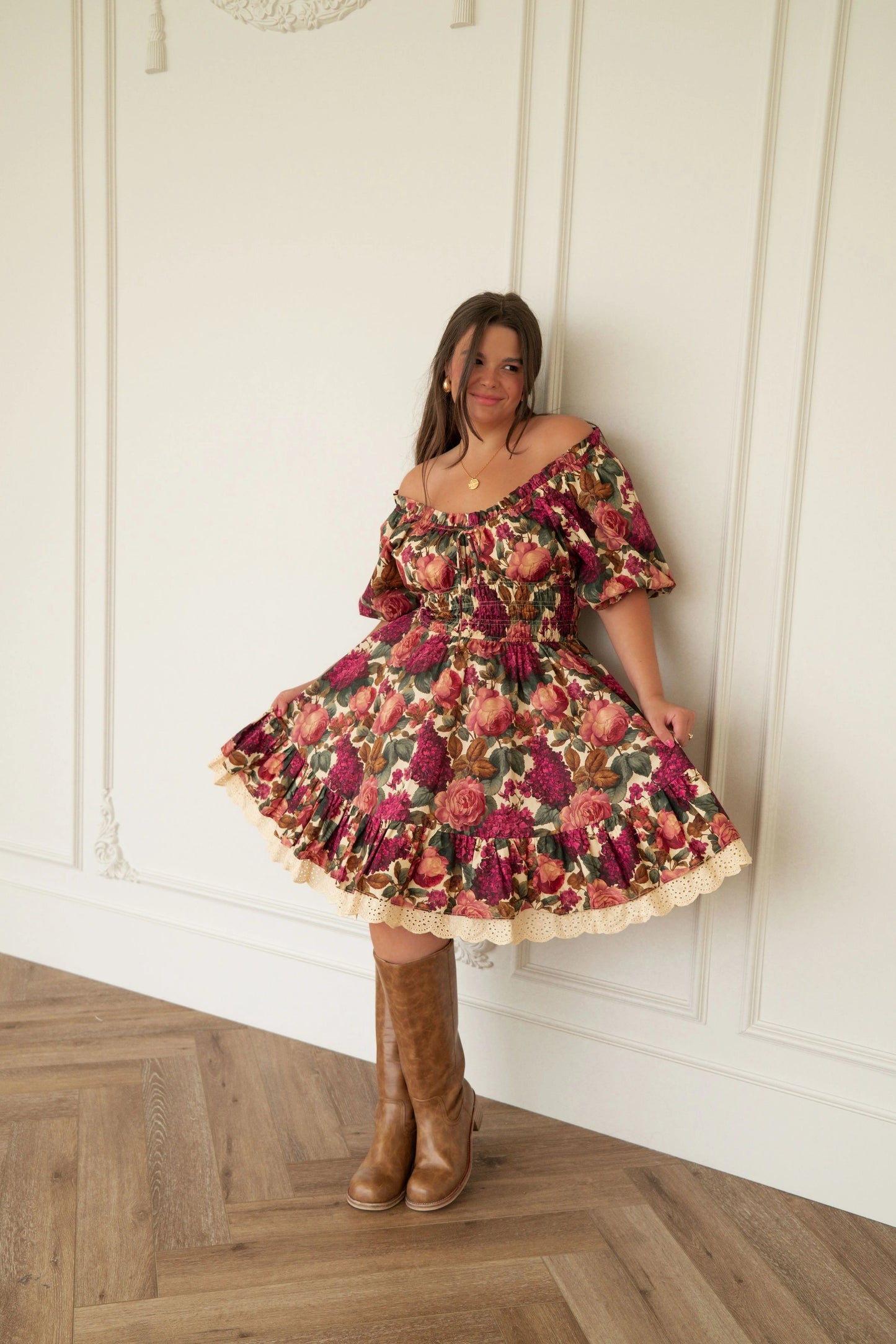 The image shows a woman standing in front of a white wall wearing a dress and brown boots. She has a confident stance and her hair is styled in a sleek bob. Her dress is a light color with a pattern