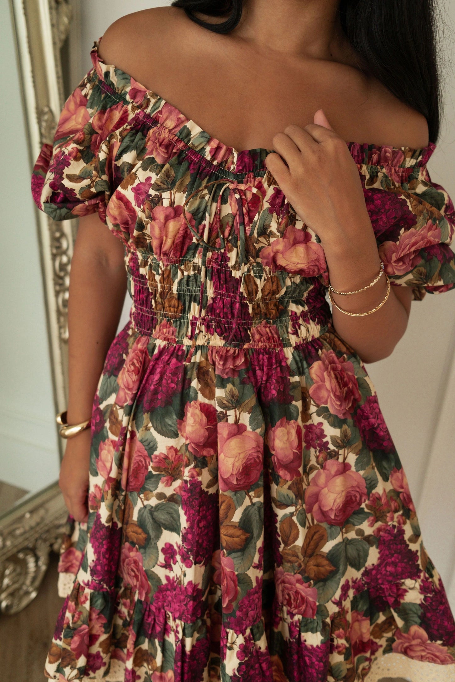 The image shows a woman standing in front of a mirror wearing a beautiful floral dress. The dress is a vibrant mix of colors, including pink, yellow, and blue, and has a delicate floral pattern. The