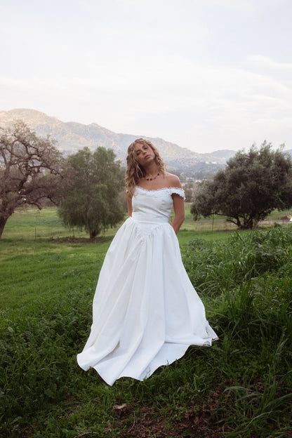 Woman in white off-the-shoulder wedding dress standing in a grassy field with trees and mountains in the background.