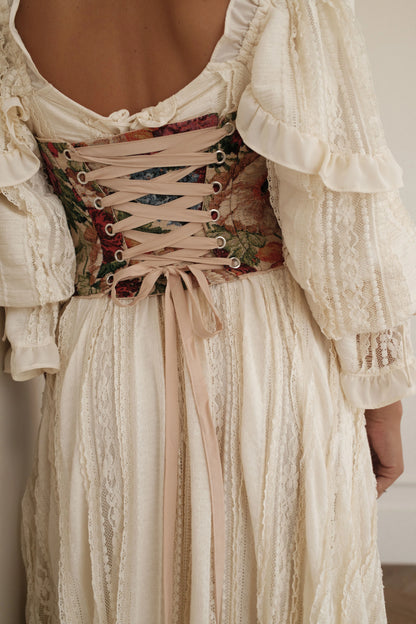 The image shows a woman wearing a white dress with a lace overlay and a corset, standing in front of a wall. The dress is made of a light, airy fabric and has a delicate lace trim along the neckline