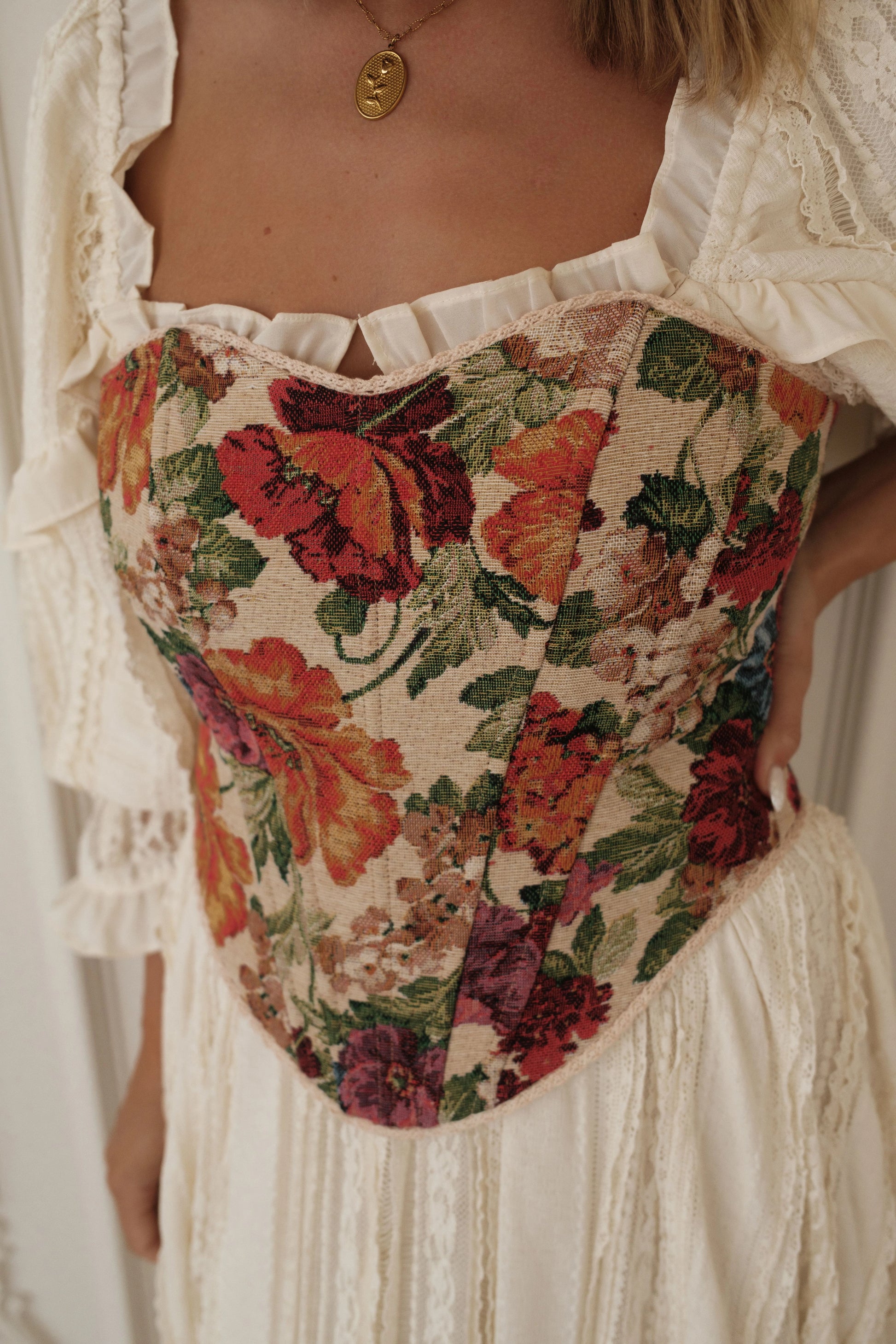 The image shows a woman wearing a white dress with a floral pattern and a necklace. She is standing in a relaxed pose, her arms crossed and her head slightly tilted to the side. Her hair is pulled
