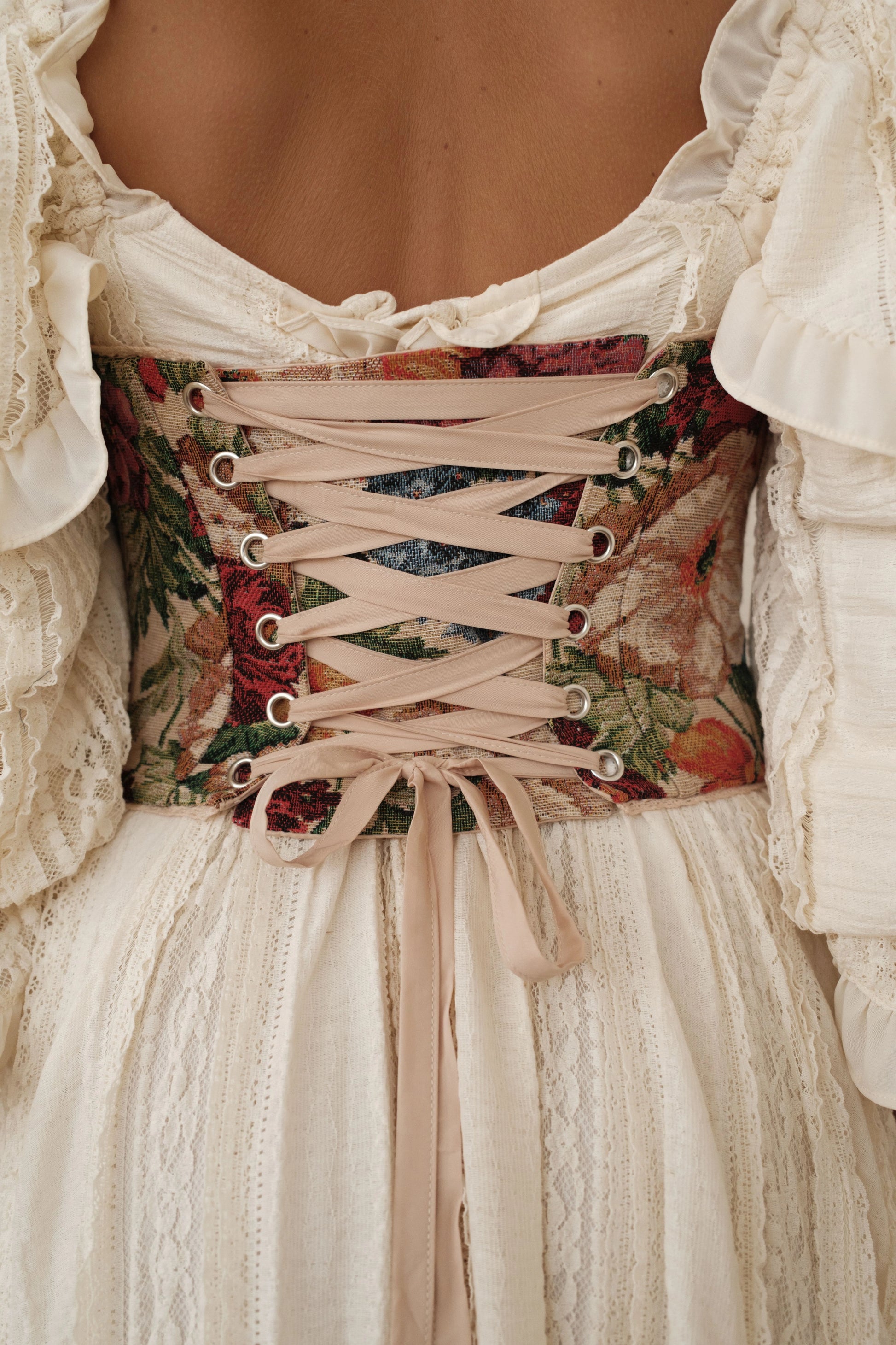 The image shows a woman wearing a white dress with a floral pattern on it. The dress is made of a light fabric and has a delicate lace-up front. The pattern is composed of various shades of pink