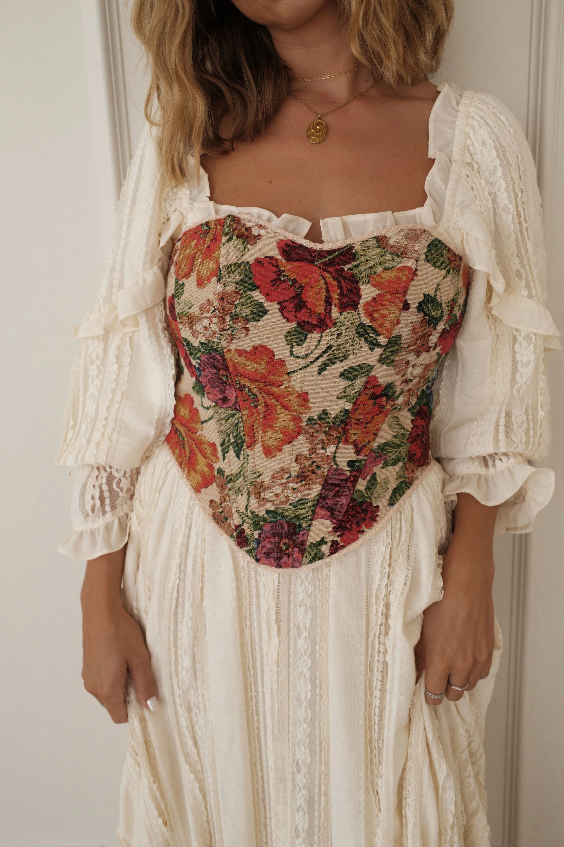 The image shows a woman standing in front of a white door wearing a white dress with a floral print. The dress has a fitted bodice and a full skirt that falls to the floor. The woman has her hands