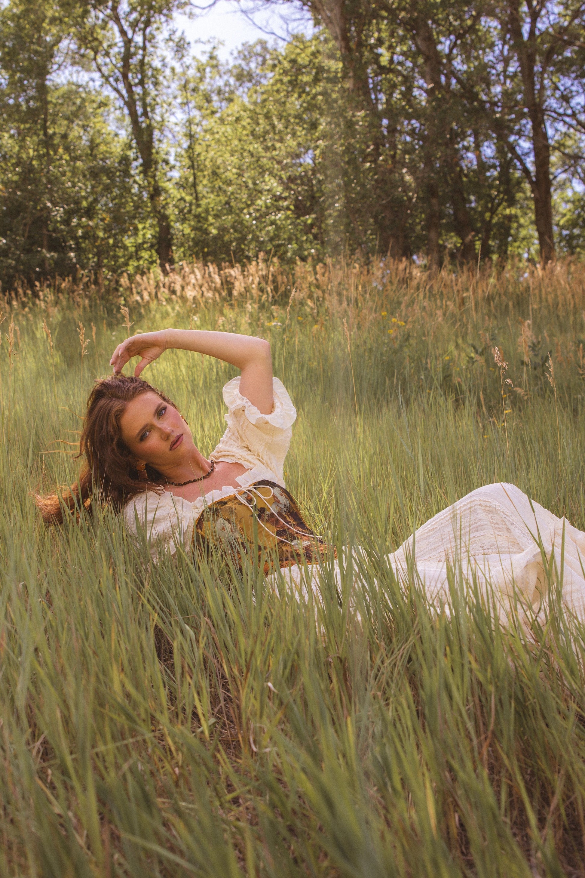 The image shows a woman in a white dress laying in a field of tall grass, surrounded by trees and a clear blue sky.