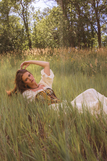 The image shows a woman in a white dress laying in a field of tall grass, surrounded by trees and a clear blue sky.