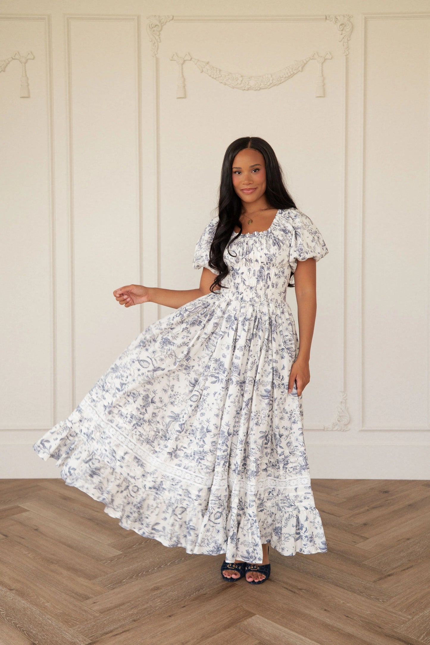 The image shows a woman standing in front of a white wall wearing a white and black dress. She is standing on the floor, with a wall behind her.