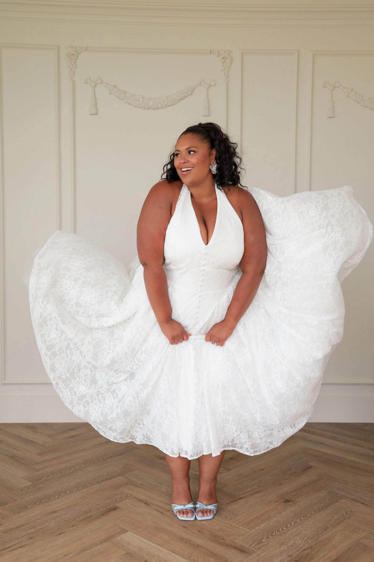 The image shows a woman in a white dress standing on a wooden floor, with a wall in the background. She is wearing a white frock, giving her a unique and stylish look.