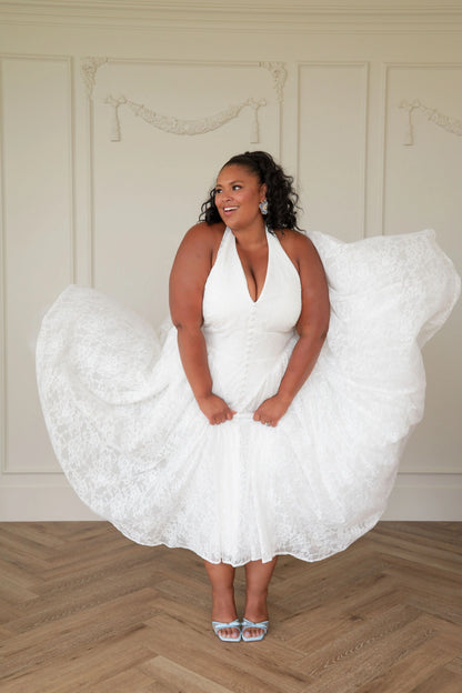 The image shows a woman in a white dress standing on a wooden floor, with a wall in the background. She is wearing a white frock, giving her a unique and stylish look.