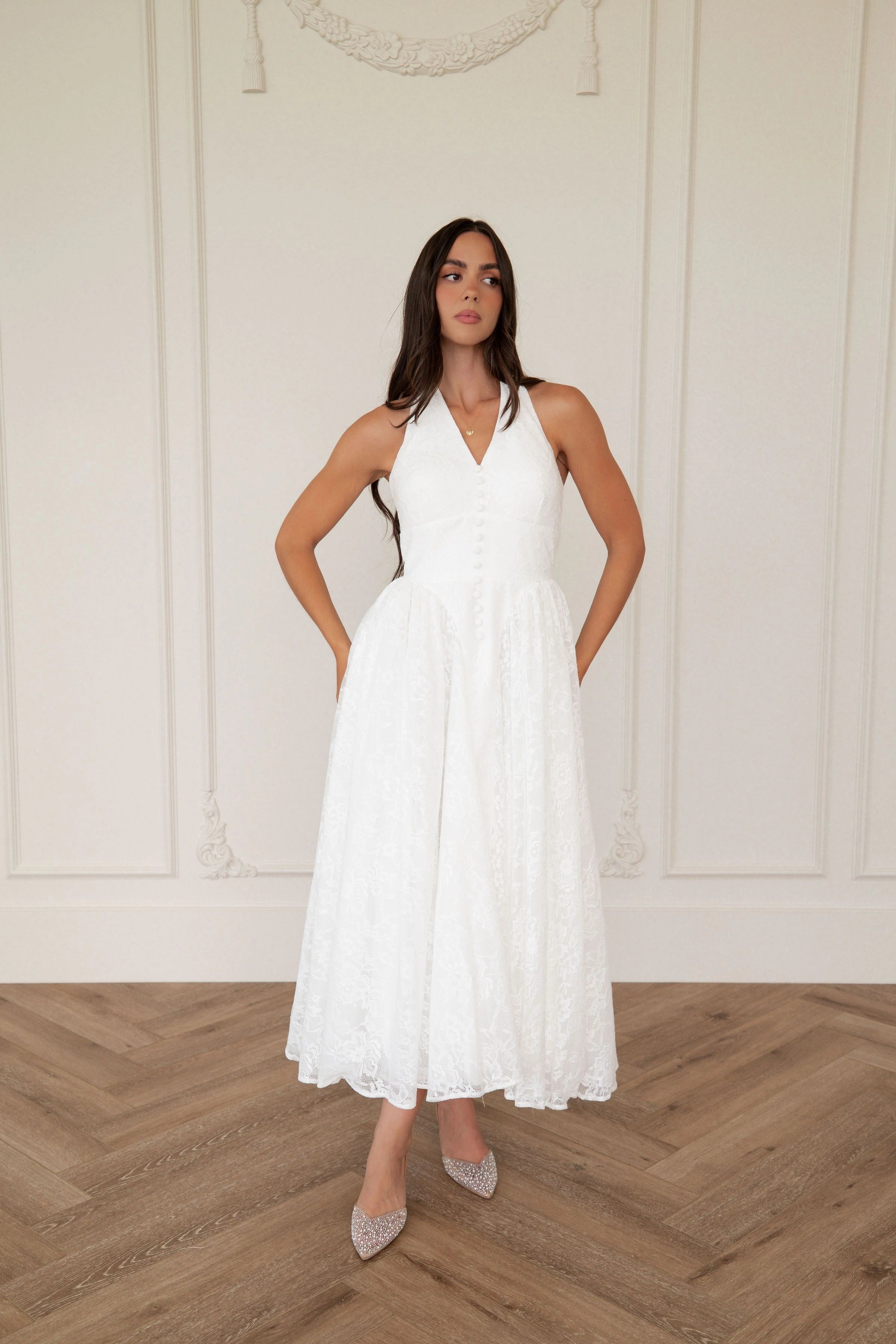 The image shows a woman standing in front of a white wall wearing a white dress. The dress is a wedding dress with a fitted bodice and a full skirt that falls to the floor. The woman has her hands on