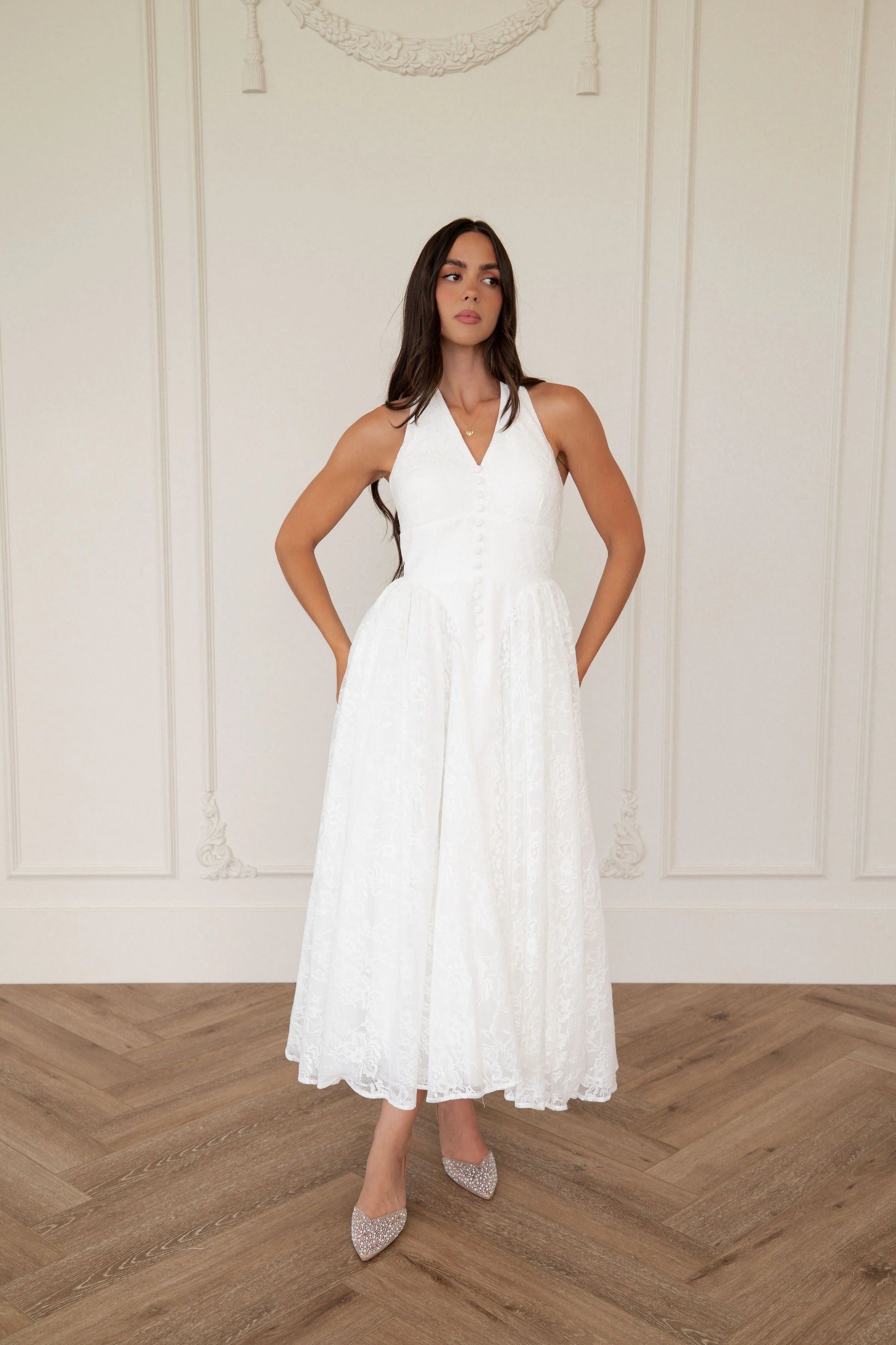 The image shows a woman standing in front of a white wall wearing a white dress. The dress is a wedding dress with a fitted bodice and a full skirt that falls to the floor. The woman has her hands on