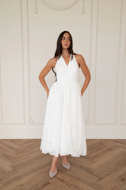 The image shows a woman standing in front of a white wall wearing a white dress. The dress is a wedding dress with a fitted bodice and a full skirt that falls to the floor. The woman has her hands on