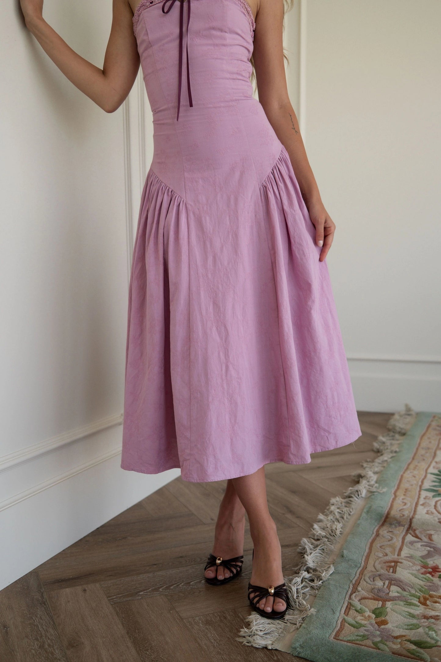 The image shows a woman standing in front of a white wall wearing a pink dress. The dress is a light pink color and has a fitted bodice with a full skirt that falls to the floor. The woman is wearing