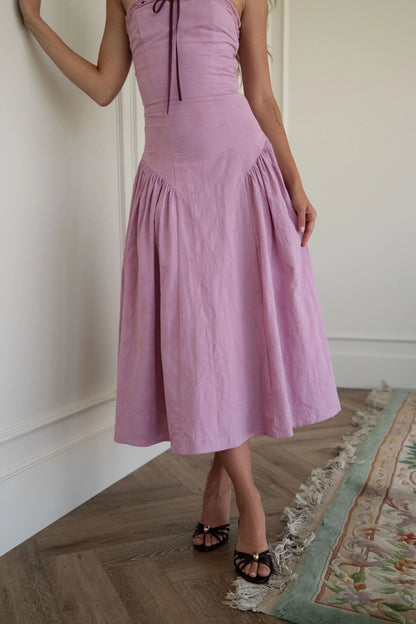 The image shows a woman standing in front of a white wall wearing a pink dress. The dress is a light pink color and has a fitted bodice with a full skirt that falls to the floor. The woman is wearing
