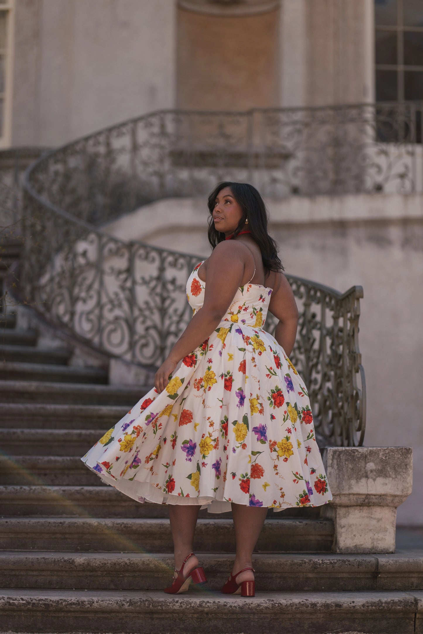 The image shows a woman in a white dress standing on the steps of a building, with a railing and a wall in the background. She is wearing a fashionable dress, likely from the latest fashion trends of