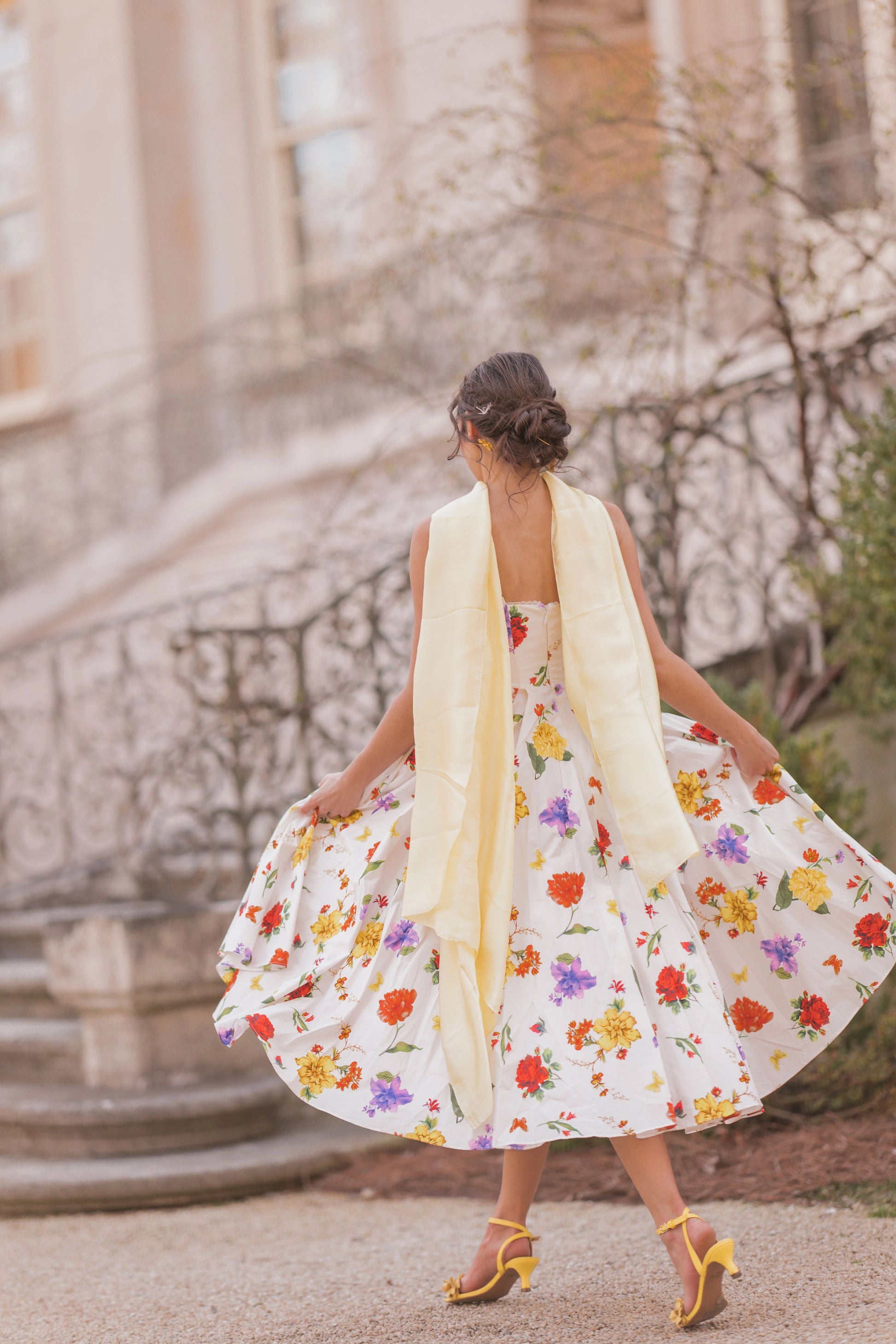 The image shows a woman in a white dress with a yellow scarf walking down a street, with a building in the background. She is wearing a white frock with a floral pattern, and the image has been