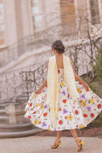 The image shows a woman in a white dress with a yellow scarf walking down a street, with a building in the background. She is wearing a white frock with a floral pattern, and the image has been