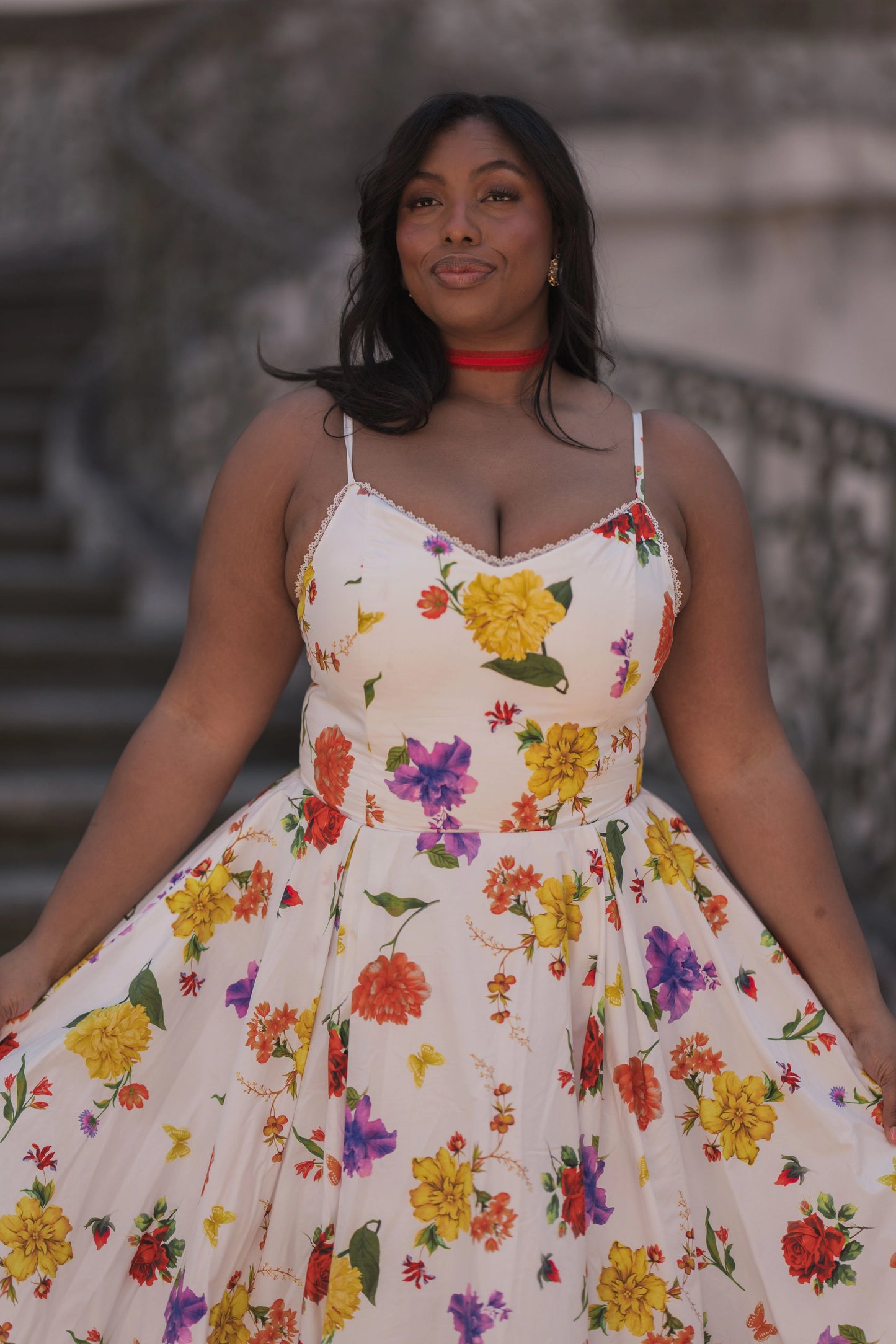 The image shows a woman in a white dress with yellow and red flowers on it, standing in front of a wall. She is wearing a red necklace and has a confident stance, as if she is ready to take on the