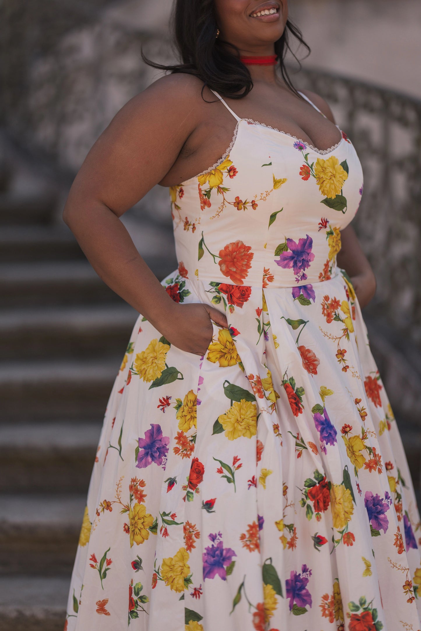 The image shows a woman in a white dress standing on a set of stairs, with a wall in the background. She is wearing a long, flowing dress with a high neckline and long sleeves, and her hair is pulled