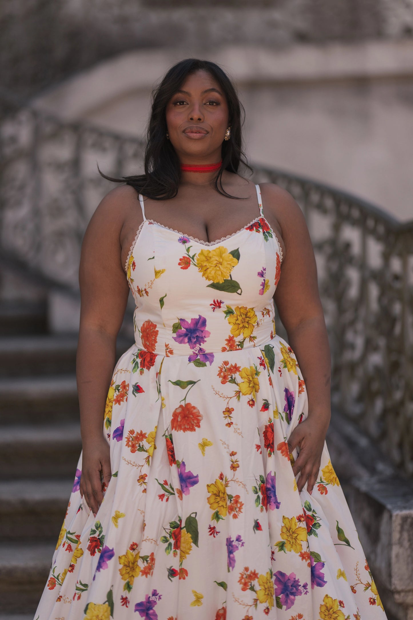 The image shows a woman in a white dress standing on a set of stairs, with a blurred background. She has a human face and is wearing a dress with a pattern of flowers.