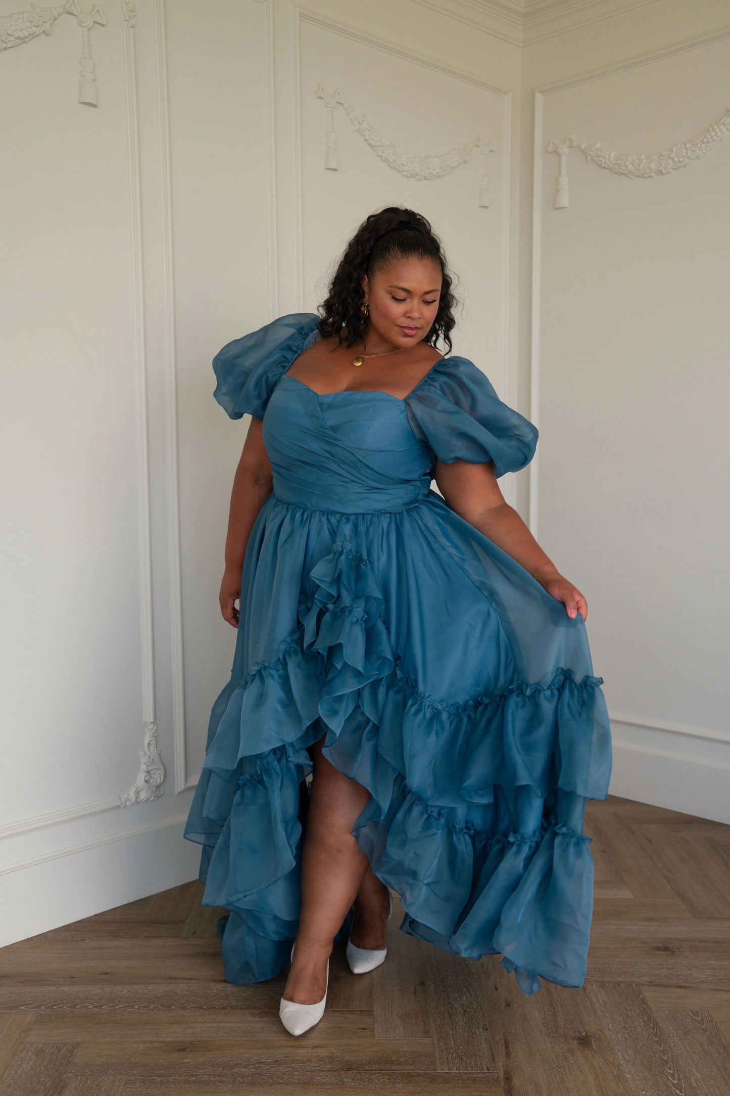 The image shows a woman standing in front of a white wall wearing a blue dress with ruffles. The dress is a deep blue color and has a fitted bodice with a full skirt that falls to the floor. The
