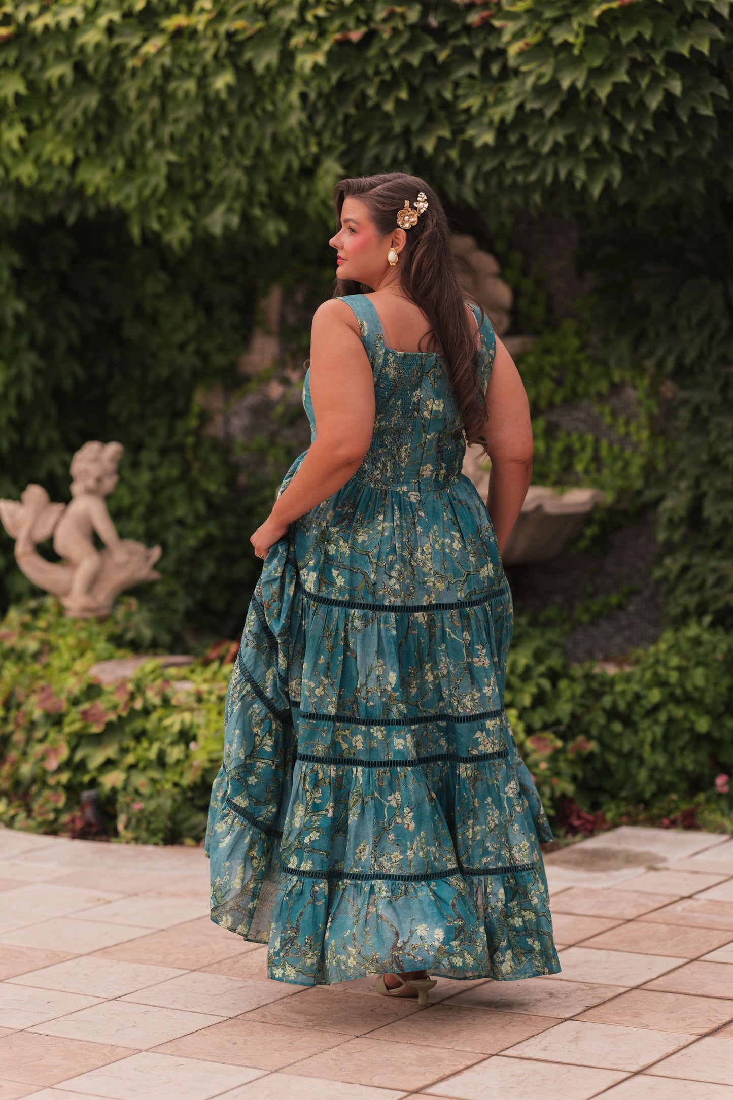 Woman in teal floral maxi dress with ruffles and gold accessories stands on beige tiles facing away from camera in garden.