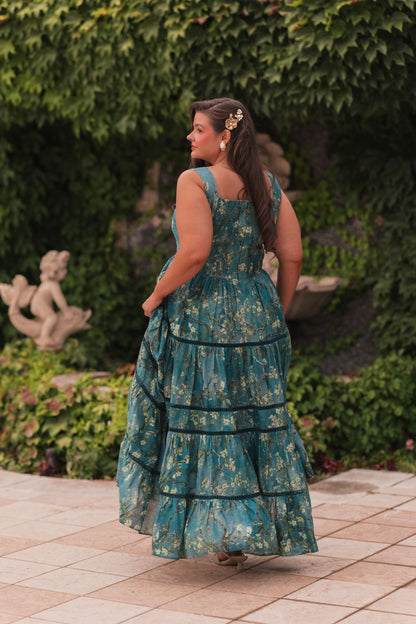 Woman in teal floral maxi dress with ruffles and gold accessories stands on beige tiles facing away from camera in garden.