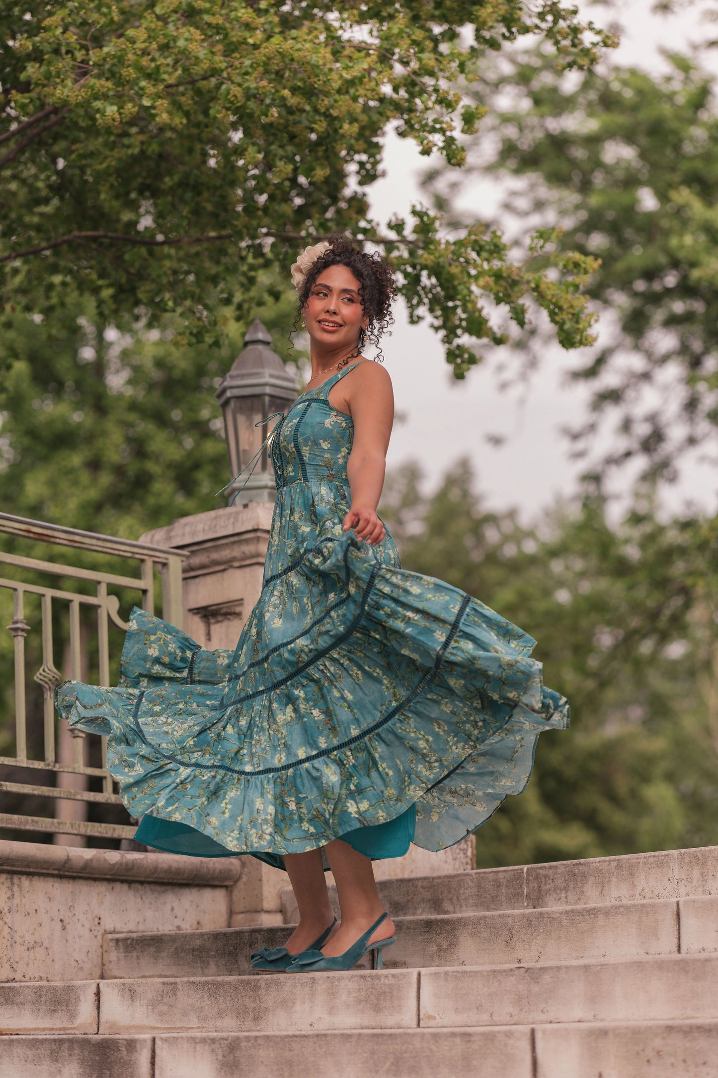 Woman in teal floral dress and flower crown standing on stone steps, smiling at camera with dress flowing outward.