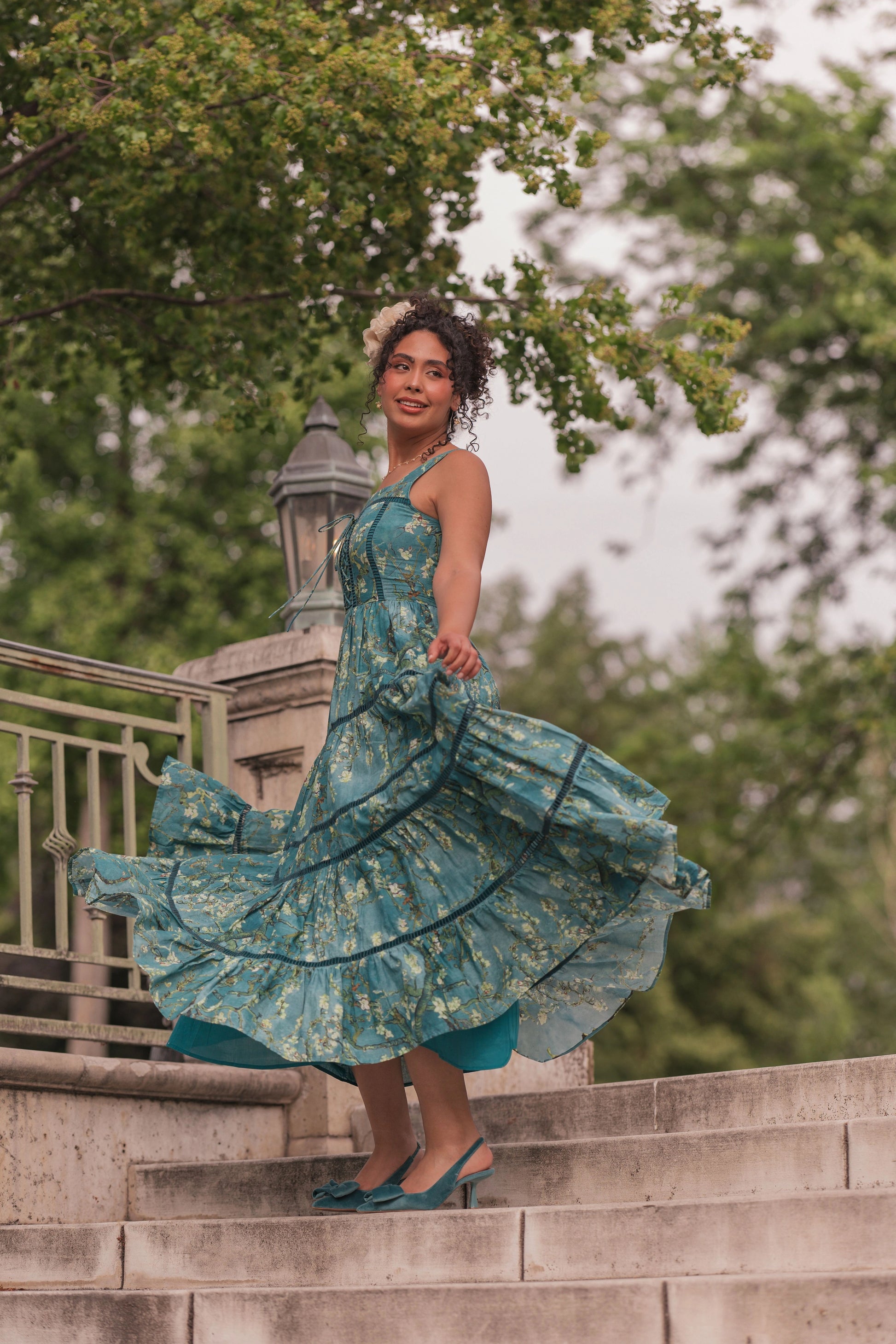 Woman in teal floral dress and flower crown standing on stone steps, smiling at camera with dress flowing outward.
