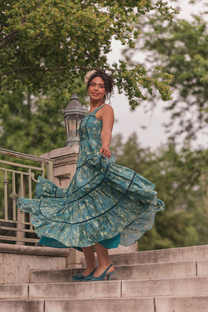 Woman in teal floral dress and flower crown standing on stone steps, smiling at camera with dress flowing outward.